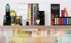 Eine Sammlung von Künstlerbedarfsprodukten der Marke Blockx, darunter Farbtuben, Aquarellfarbpaletten, Pinsel und Pastellkreide. Verschiedene leuchtende Farben sind geordnet auf einem Regal über einem bunten Mosaikhintergrund ausgestellt.
