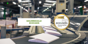 Eine Industrieanlage von Quo Vadis mit einem Förderband, auf dem offene Kalendereinlagen transportiert werden. Der Textüberzug lautet „Goldmedaille EcoVadis“. Ein Logo daneben lautet „Gold 2023 EcoVadis Sustainability Rating“. Im Hintergrund sind Produktionsmaschinen zu sehen.
