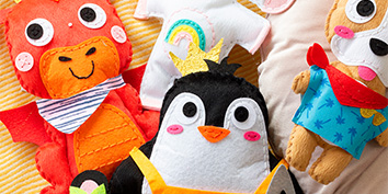 Eine Sammlung von genähten Plüschtieren, darunter ein roter Drache, ein Pinguin mit goldener Krone und ein brauner Hund in blauem Outfit, sind auf einer gestreiften gelben Decke angeordnet. Die Spielzeuge zeichnen sich durch leuchtende Farben und detaillierte Nähte aus.