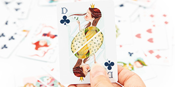 Eine Person hält eine Spielkarte mit der Kreuz-Dame in einem einzigartigen Design, das eine skurrile, cartoonhafte Figur zeigt. Andere Karten sind im Hintergrund verstreut.