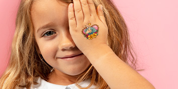 Ein junges Mädchen mit langen blonden Haaren lächelt und bedeckt ein Auge mit ihrer Hand, auf der sich ein buntes temporäres Tattoo von My Mandarine eines blauen Elefanten mit Blumen befindet. Der Hintergrund ist rosa.