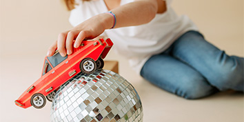Eine Kind in weißem Shirt und Jeans spielt mit einem roten Spielzeugauto auf einer reflektierenden Discokugel.