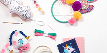 Kreativset von My Mandarine mit genähtem Einhorn, Stoffpuppe zum Ausmalen, Farbtöpfen, Stickgarn, einem rosa Stoffquadrat, einem Traumfänger und einem hölzerner Stickrahmen auf einer weißen Oberfläche.