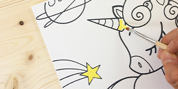 Eine Person malt mit einem Pinsel eine Illustration eines Einhorns und fügt dem Horn gelbe Farbe hinzu. Die Zeichnung enthält außerdem einen Stern, einen Planeten und eine Sternschnuppe vor einem Holzhintergrund.