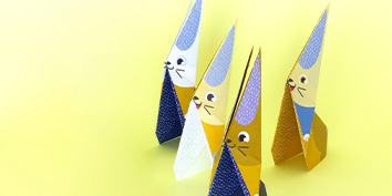 Vier Origami-Figuren von My Mandarine in Form von Kaninchen auf gelbem Hintergrund. Jedes Kaninchen hat ein farbenfrohes Design mit unterschiedlichen Mustern, darunter Blau- und Gelbtöne. Sie sind leicht versetzt angeordnet und blicken in die gleiche Richtung.