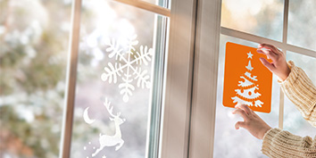Ein Kind in einem cremefarbenen Pullover hält eine orangefarbene Schablone mit einem Weihnachtsbaummotiv von My Mandarine an ein Fenster. Das Fenster ist mit weißen Schneeflocken- und Rentieraufklebern dekoriert. Draußen suggeriert verschwommenes Laub eine winterliche Kulisse.