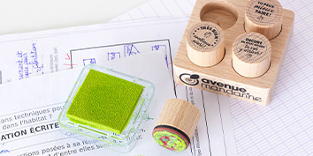 Ein Stapel mit Klassenarbeiten mit einem grünen Stempelkissen, einem kleinen runden Stempel und einem Holzblock mit drei Stempelschlitzen von My Mandarine. Die Stempel zeigen Sätze auf Französisch und sind auf einer hellen Unterlage angeordnet.