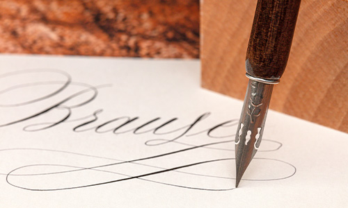Eine Nahaufnahme eines Kalligrafiestifts, der das Wort „Brause“ auf weißes Papier schreibt. Der Stift hat eine Metallspitze und einen Griff aus dunklem Holz. Die Schrift ist elegant und fließend, mit komplizierten Schleifen und dünnen Strichlinien.