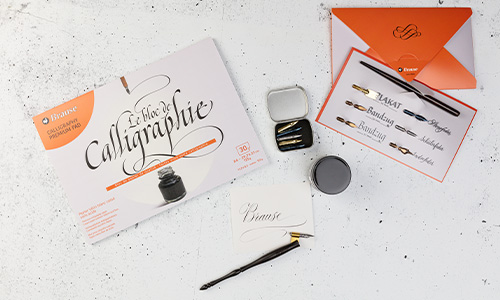 Kalligraphie-Set auf weißem Untergrund, bestehend aus Kalligraphieheft, Tintenfass, schwarzem Stift mit Feder, zusätzlichen Federn in einer Dose, Übungsblättern und orange-weißem Umschlag. Das Heft trägt die Aufschrift „Kalligraphie“.