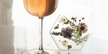 Ein Glas Roséwein steht auf einer reflektierenden Oberfläche neben einem sechseckigen Untersetzer mit gepressten Blumen und Goldflocken. Weiches, natürliches Licht fällt durch ein Fenster im Hintergrund.