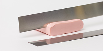 Auf einer weißen Oberfläche liegt ein Block rosa Modelliermasse von Cernit, darauf balanciert eine Modellierklinge aus Metall, unter der sich die Reflexionen spiegeln.