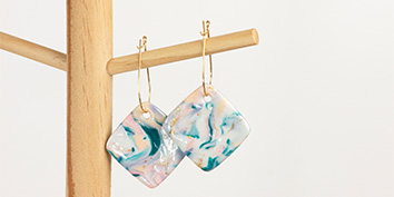 Hängende Ohrringe aus Modelliermasse von Cernit mit quadratischen Harzanhängern mit blau-weiß- und pfirsichfarbenem Marmormuster, befestigt an einem Holzständer.