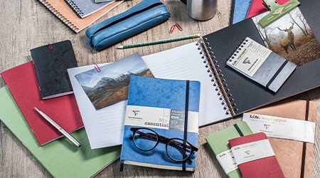 Verschiedene Schreibwaren auf einem Holztisch, darunter Notizbücher, ein roter und ein schwarzer Stift, ein Federmäppchen, Fotos und ein Bleistift. Eine Brille liegt auf einem blauen Notizbuch. Es handelt sich um die Kollektion Age Bag von Clairefontaine.
