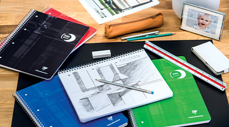 Ein Schreibtisch mit bunten Collegeböcken mit dot grid von Clairefontaine, einem Federmäppchen, Bleistiften, einem Radiergummi, Lineal und einem offenen Collegeblock mit einer Gebäudezeichnung. Im Hintergrund ist ein gerahmtes Foto eines lächelnden Kindes zu sehen. Die Umgebung ist hell und organisiert.