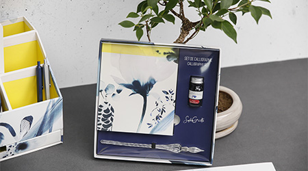 Ein Kalligraphie-Set mit einer Glasfeder, einem Tintenfass und Papier steht auf einer grauen Oberfläche. Die Box zeigt ein abstraktes Blumenmuster. Im Hintergrund sind eine kleine Topfpflanze und ein weißer Organizer mit gelben Gegenständen zu sehen. Es handelt sich um die Kollektion Cyanotype von Clairefontaine.