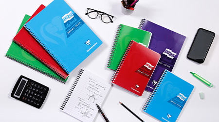 Eine Sammlung bunter Spiralblöcke, ein Smartphone, ein Taschenrechner, eine Brille, ein Stifthalter mit Stiften und ein Textmarker sind auf einer weißen Oberfläche angeordnet. Ein offenes Notizbuch zeigt handschriftliche Notizen und ein Diagramm. Es handelt sich um die Kollektion Europa von Clairefontaine.