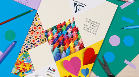 Eine Auswahl an farbenfrohen Bastelutensilien auf einem Tisch, darunter Papierausschnitte, Scheren, Marker und ein Block mit buntem Papier von Clairefontaine mit einem Umschlag mit verschiedenen Buntstiften und Mustern.