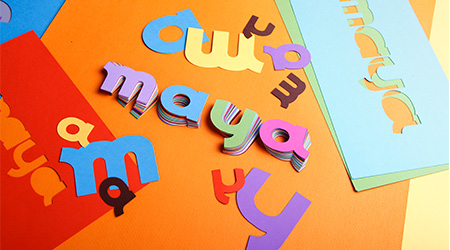 Ausgeschnittene, bunte Buchstaben aus Papier ergeben das Wort „Maya“ und sind auf einer orangefarbenen Oberfläche gestapelt, umgeben von weiteren verstreuten Buchstaben und farbigen Papierbögen in Rot, Blau und Grün. Es handelt sich um Maya-Papier von Clairefontaine.