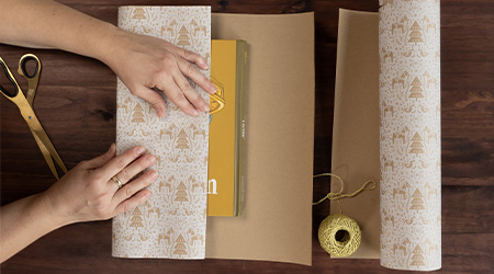 Frau packt Bücher mit Kraft-Geschenkpapier mit Tannenbäumen von Clairefontaine ein. Auf braunem Holztisch, auf dem auch eine goldene Schere und ein Bast-Geschenkband liegt.