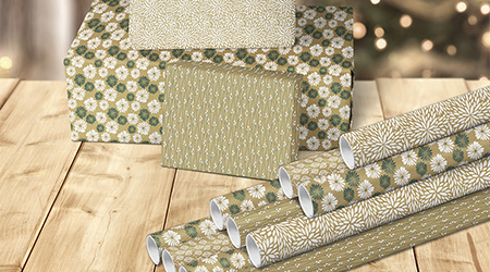 Grün-braune Geschenkpapierrollen von Clairefontaine und damit eingepackte rechteckige Geschenke in 3 verschiedenen Größen. Alles auf hellbraunem Holztisch. Im Hintergrund leuchten unscharf die Lichter vom Tannenbaum.