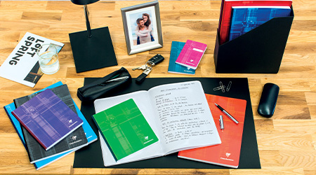 Ein Holzschreibtisch mit verschiedenen Gegenständen: offene Notizbücher und Kladden von Clairefontaine, farbige Ordner, eine kleine Lampe, ein gerahmtes Foto, Autoschlüssel, ein Smartphone, Stifte und eine Maus. Ein Glas Wasser steht neben einer Zeitschrift, die teilweise sichtbar ist.