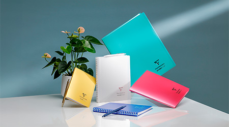 Farbenfrohe Notizbücher in Blau, Rosa, Gelb und Weiß sowie einem Stift auf einer weißen Oberfläche. Eine grüne Topfpflanze sorgt für eine natürliche Note und der Hintergrund ist in einem satten Blaugrün gehalten, durch das ein Lichtstrahl scheint. Es handelt sich um die Kollektion Koverbook von Clairefontaine.