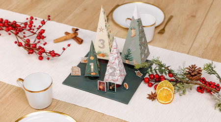 Dekorative Papierbäume mit Feiertagsmustern von Clairefontaine sind auf einem Tischläufer arrangiert. Umgeben von roten Beeren, Tannenzapfen und getrockneten Orangenscheiben steht die Szene auf einem Holztisch mit goldumrandeten Tassen und Besteck.