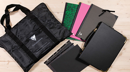 Eine schwarze Portfoliotasche, umgeben von mehreren Zeichenmapppen und Mappen von Clairefontaine in verschiedenen Farben und Mustern, darunter Rosa, Grün und Schwarz. Sie liegen alle auf einer hellen Holzoberfläche.