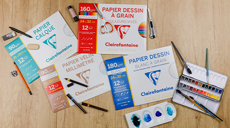 Auf einer Holzoberfläche sind Künstlerutensilien angeordnet, darunter Packungen mit Clairefontaine-Zeichenpapier in verschiedenen Texturen und Größen. Um sie herum befinden sich Pinsel, Bleistifte, Aquarellnäpfe und eine Palette mit Blautönen.