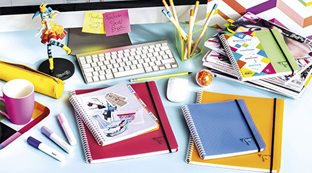 Schreibtisch mit bunten Notizbüchern von Clairefontaine, einer Tastatur, Stiften, Bleistiften, Haftnotizen und einer Spielzeugfigur. Die Notizbücher haben ein geriffeltes Muster. Auf der Haftnotiz steht „Fashion School Goals“. Heller und organisierter Arbeitsplatz.