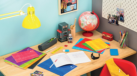 Ein bunter Schreibtisch mit Notizbüchern und Schulheften von Clairefontaine, einem Globus, einer Lampe, einem kleinen Roboter und einem Stifteköcher. Auf dem Tisch liegen Fotos und ein Spielzeugbus. Die Wand zieren ein Comic-Poster und eine Stecktafel mit Fotos.