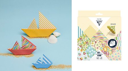 Bunte Schiffchen aus Origamipapier von Clairefontaine auf hellblauem Hintergrund, mit Sand, so dass es aussieht als würden sie am Strand stehen. Rechts daneben die Verpackung der Papiere.