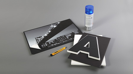 Auf einer grauen Oberfläche sind ein Schwarzweißfoto einer Brücke und der Skyline einer Stadt, ein Universalmesser und ein schwarzer Ausschnitt des Buchstabens „A“ angeordnet. Hinter den Gegenständen befindet sich eine Dose Sprühfarbe mit blauem Deckel. Es handelt sich um Pappe, Kartons und Schaumkartons von Clairefontaine.