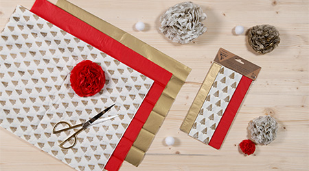 Weihnachtliches Seidenpapier in gold, rot und weiß mit Tannenbäumen von Clairefontaine auf hellbraunem Holztisch liegend. Zusammen mit einer Schere und Kreppblume. Daneben die 3 Seidenblätter in der Verpackung. Blick von oben.