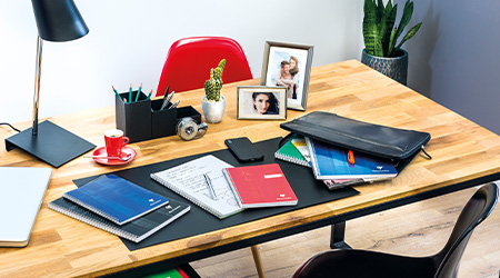Ein Holzschreibtisch mit einer schwarzen Schreibtischunterlage, Notizbüchern von Clairefontaine, einem Laptop, einem Smartphone und einer roten Kaffeetasse. Auf dem Schreibtisch stehen eine Lampe, ein Bleistifthalter, eine Topfpflanze und gerahmte Fotos. Im Hintergrund stehen zwei rote Stühle und eine größere Topfpflanze.