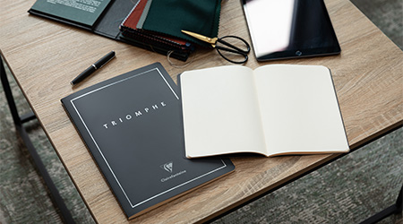 Ein Holzschreibtisch mit einem schwarzen Notizbuch mit der Aufschrift „Triomphe“, einem offenen, leeren weißen Notizbuch, einem Stift, einer Schere, Stoffmustern und einem Tablet. Die Umgebung lässt an einen Arbeits- oder Lernbereich denken. Es handelt sich um die Kollektion Triomphe von Clairefontaine.