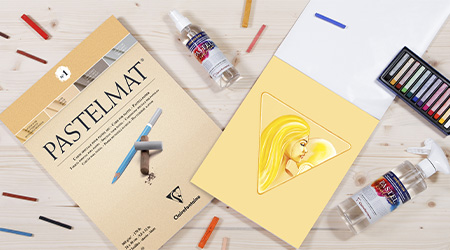 Auf einem Tisch liegen Künstlerutensilien, darunter ein Pastelmat-Block und Fixierspray von Clairefontaine sowie Pastellstifte und eine Zeichnung mit einer Figur mit gelben Haaren auf einem dreieckigen gelben Blatt. Die Szene erinnert an eine Pastellkunst-Ausstellung.
