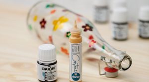 Auf einer Holzoberfläche liegt eine klare Glasflasche, die mit bunten Blumenmotiven verziert ist. Bemalt wurde sie mit Glasmalfarbe von Darwi. Im Vordergrund sind eine offene Tube mit goldener Farbe und eine kleine Flasche mit der Aufschrift „Darwi Glass“ zu sehen. Im Hintergrund sind weitere Farbflaschen verschwommen.