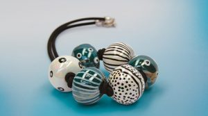 Ein Armband mit großen, dekorativen Perlen auf einer schwarzen Kordel. Die Perlen sind mit verschiedenen Mustern verziert, darunter Streifen, Tupfen und abstrakte Designs in den Farbtönen Blaugrün, Schwarz und Weiß. Gemalt mit kalter Keramikfarbe von Darwi. Der Hintergrund ist ein hellblauer Farbverlauf.