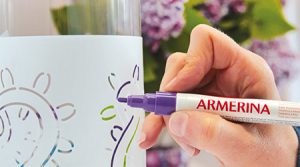 Eine Hand hält einen lilafarbenen Keramikmarker von Darwi und malt auf einer Glasoberfläche Blumenmuster, im Hintergrund sind violette Blumen verschwommen. Der Marker trägt die Aufschrift „Armerina“.