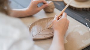Eine Person fertigt einen Tonteller aus Modelliermasse von Darwi in Blattform an und schnitzt mit einem Holzwerkzeug Adern in das Muster. Die Arbeitsfläche ist mit Papier bedeckt, im Hintergrund sind weitere Tonstücke zu sehen.