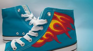 Blaue High-Top-Sneaker mit weißen Schnürsenkeln und Sohlen sowie roten und gelben Flammenmotiven an den Seiten auf hellblauem Untergrund, gemalt mit Textilmalfarbe von Darwi.