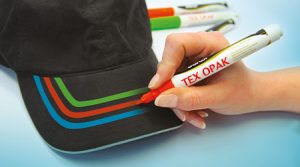Handzeichnung farbiger Streifen auf dem Schirm einer schwarzen Kappe mit einem roten Textilmarker von Darwi mit der Aufschrift „TEX OPAK“. Im Hintergrund sind weitere Stifte in Grün und Orange zu sehen.
