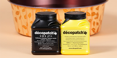 Zwei Behälter mit Klarlack mit der Aufschrift „Décopatch“ und schwarzem und gelbem Deckel stehen vor einer dekorativen Schale mit Blättermuster. Der schwarze Behälter trägt die Aufschrift „Aqua Pro“. Beide Behälter enthalten Klarlack.