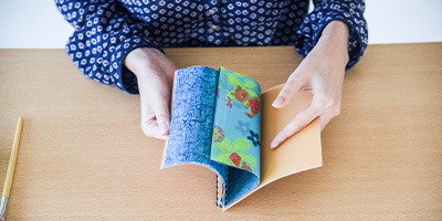 Eine Person in einem gemusterten blauen Hemd sitzt an einem Holztisch und blättert in einem bunt illustrierten Buch mit blau-grünem Einband. In dem Buch befinden sind verschiedene Blätter Décopatch-Papier. Links neben dem Buch liegt ein Bleistift auf dem Tisch.