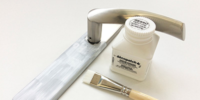Auf einer weißen Fläche liegt ein teilweise weiß lackierter Türgriff aus gebürstetem Metall. Daneben liegen ein flacher Pinsel mit Holzgriff und eine kleine weiße Flasche weißer Acrylfarbe mit der Aufschrift „Décopatch“.