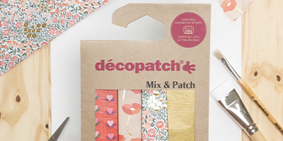 Ein Déopatch Mix & Patch-Set auf einer Holzoberfläche mit verschiedenen dekorativen Papieren in floralen und abstrakten Mustern. Pinsel liegen in der Nähe und suggerieren eine Bastel- oder Heimwerkeraktivität.