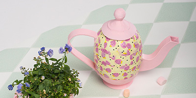 Eine rosa-gelbe dekorierte Teekanne mit Blumenmuster von Décopatch steht auf einer grün-weiß karierten Oberfläche. Daneben steht eine kleine Pflanze mit lila Blüten. Daneben liegen zwei kleine pastellfarbene Marshmallows.
