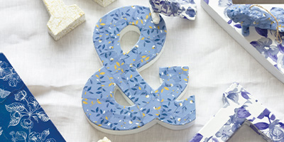Ein dekoratives Und-Zeichen mit blauem Blumenmuster und goldenen Akzenten ist von weiteren gemusterten Buchstaben auf einer hellen Stoffoberfläche umgeben. Von Décopatch.