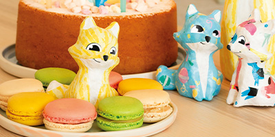 Drei bunte Fuchsfiguren von Décopatch stehen auf einem Tisch neben einem Teller mit verschiedenen Macarons in Pink, Grün, Gelb und Beige. Im Hintergrund steht eine Torte mit rosa Verzierungen. Das Ambiente wirkt verspielt und festlich.